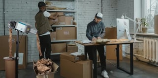 Two young men working in a fulfillment center, surrounded by boxes and a standing desk.
